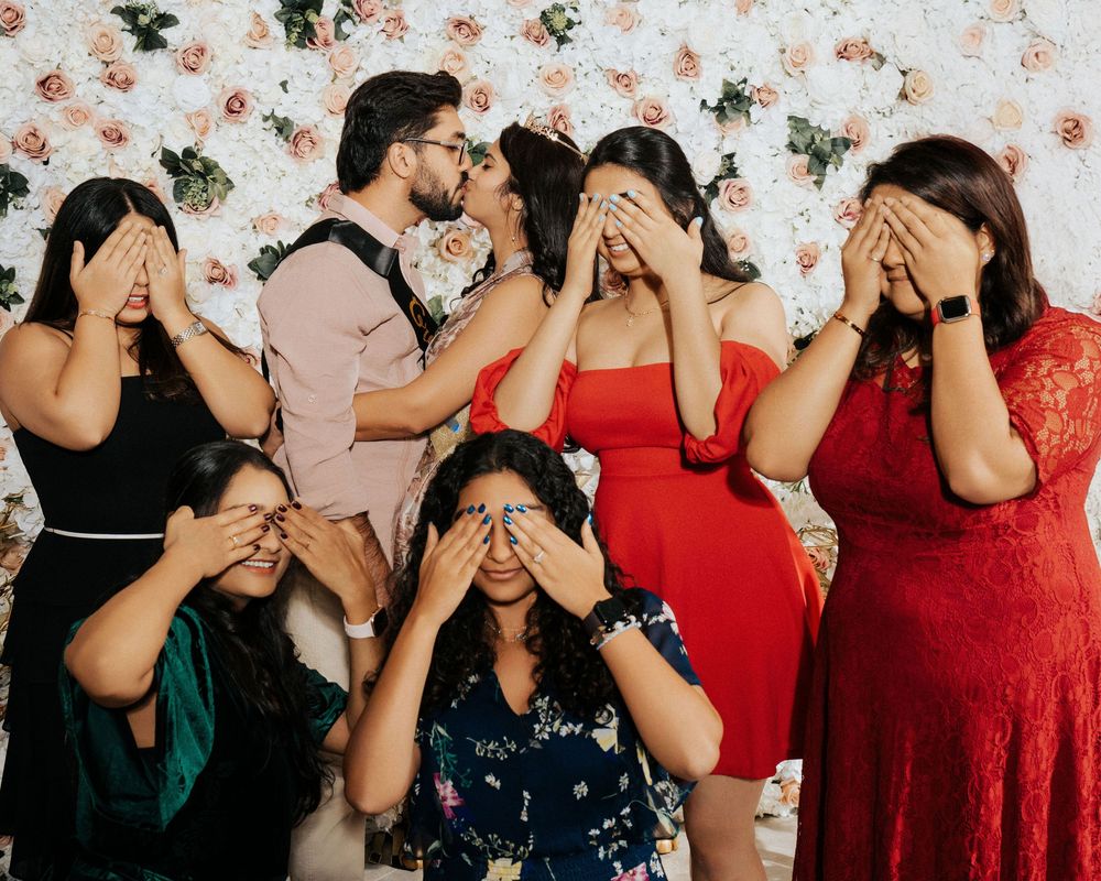 A group in formal dresses poses with hands covering their eyes against a floral wallpaper background.