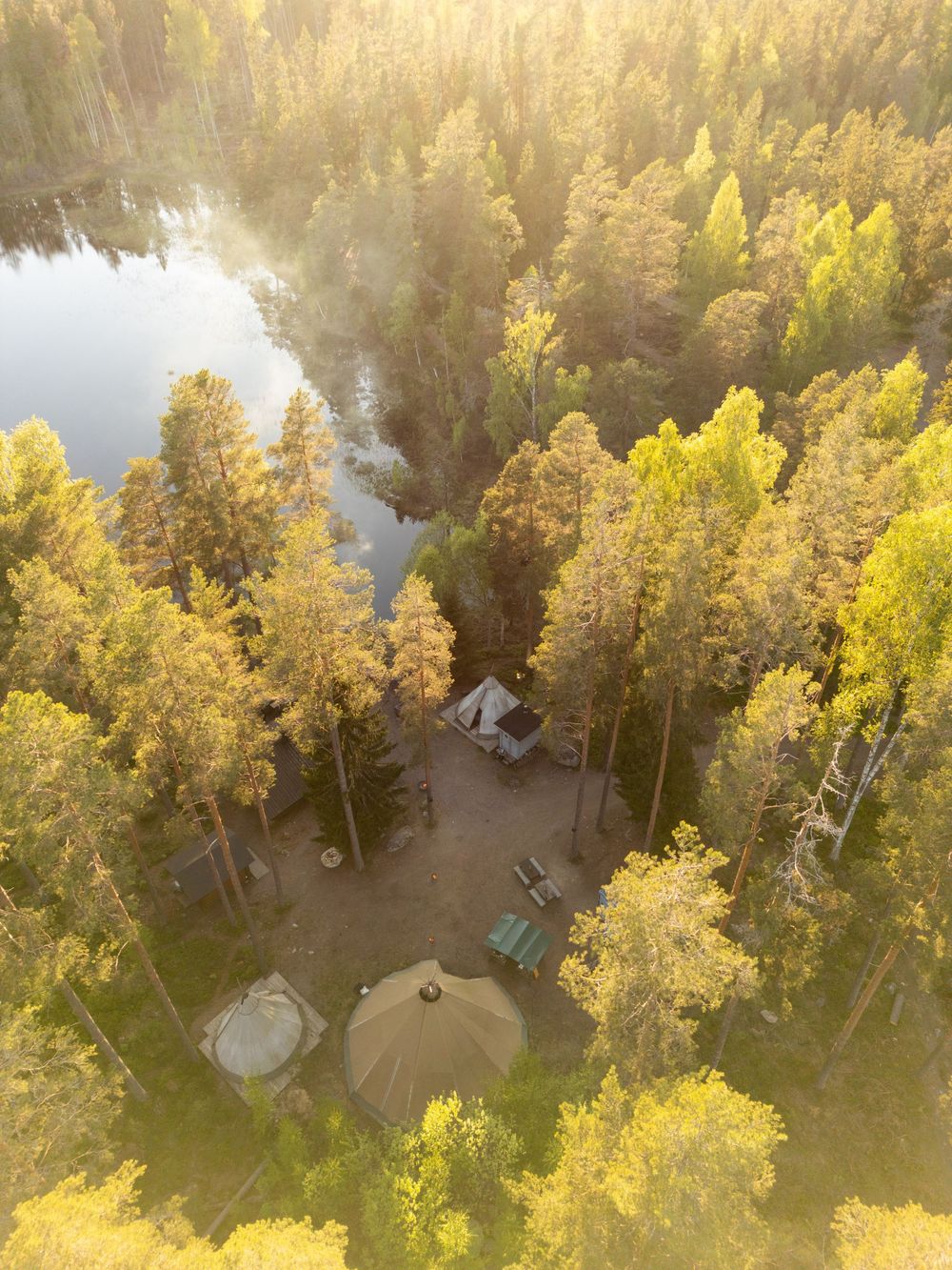 Aerial view of a remote forest campsite with a cabin and yurt nestled among pine trees beside a serene lake.