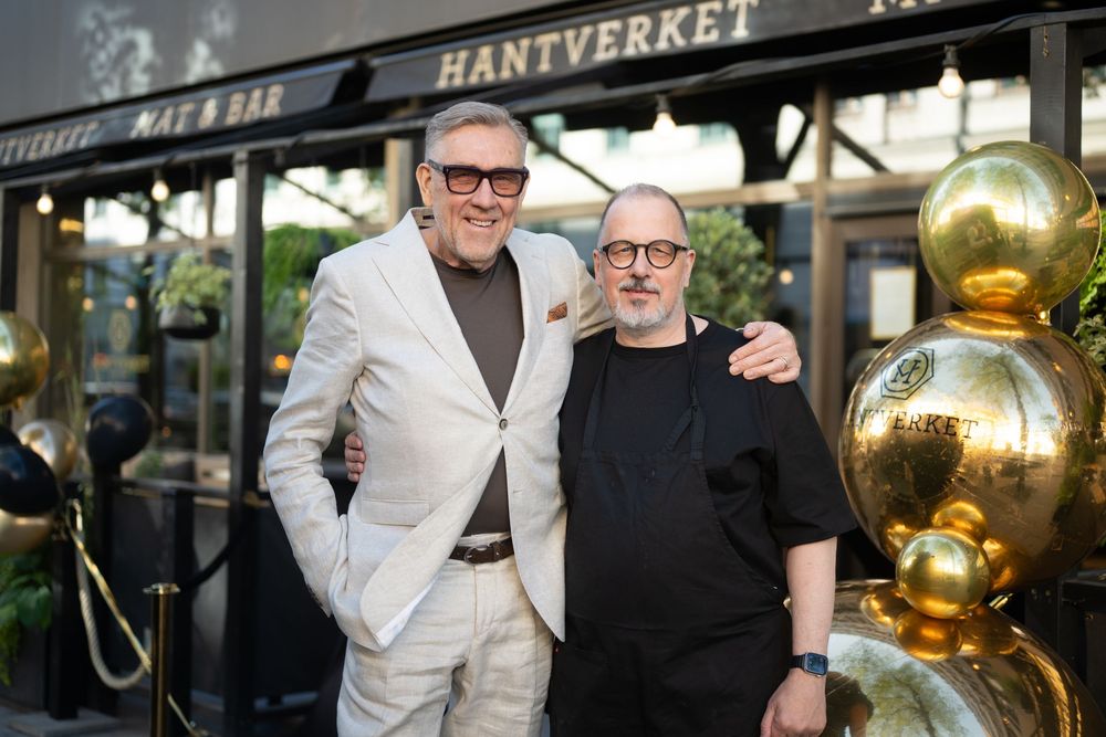 Two men pose smiling outside Hantverket restaurant, one in beige suit, one in black, with gold balloons.