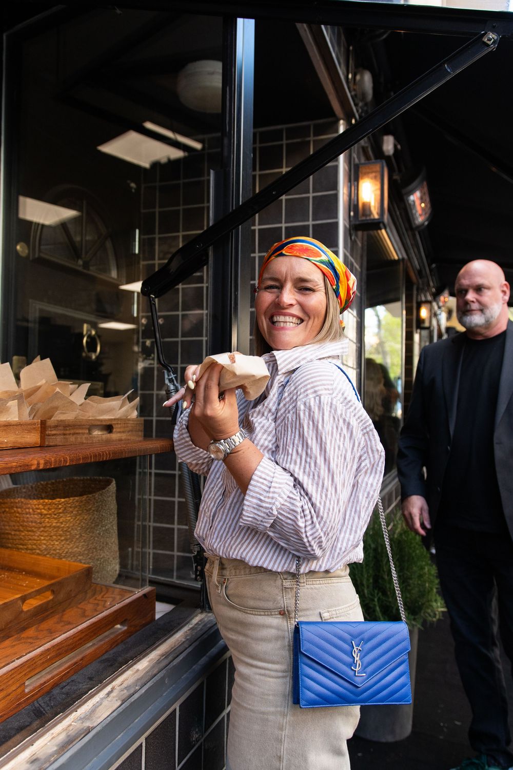 Smiling woman in striped shirt and colorful headscarf holds food, wearing blue YSL crossbody bag outside a restaurant.