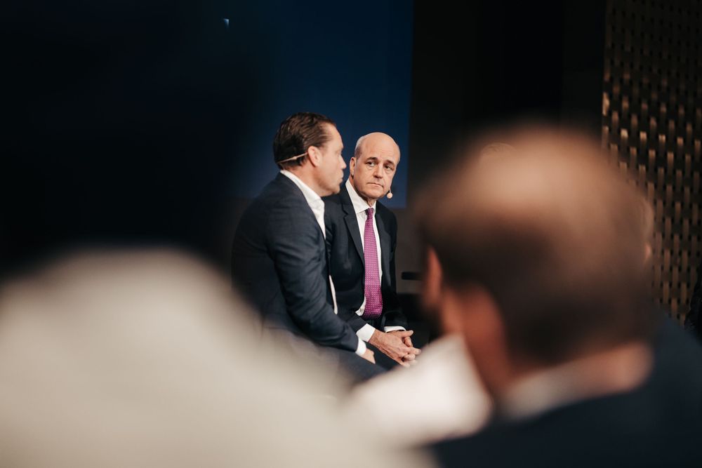 Two business professionals in suits engaged in conversation during a meeting with others visible in foreground blur.