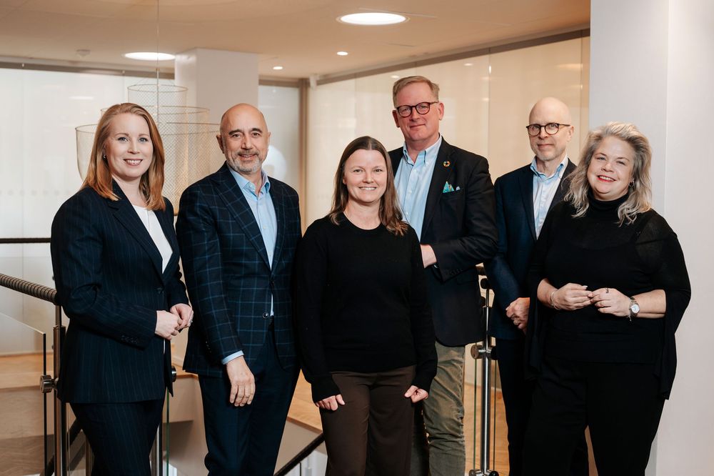 Six business professionals pose together smiling in a modern office hallway, dressed in formal attire.
