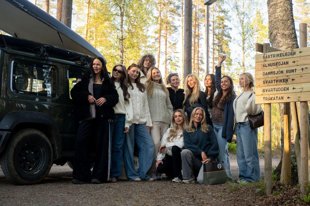 Group of young women posing together near a camper van and wooden signpost in a sunny forest setting.