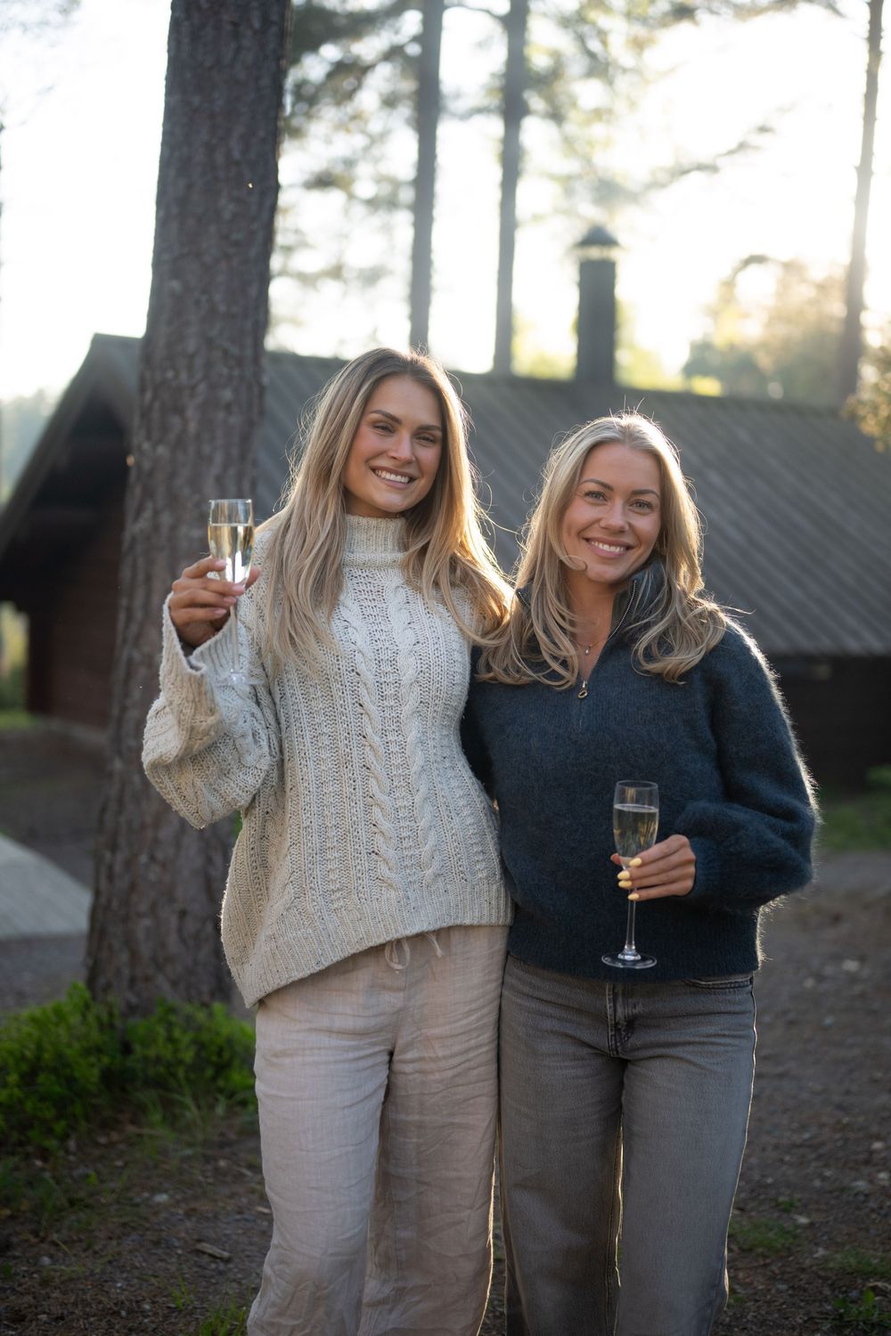 Two smiling women in cozy sweaters toasting with champagne glasses outdoors near pine trees and a cabin.