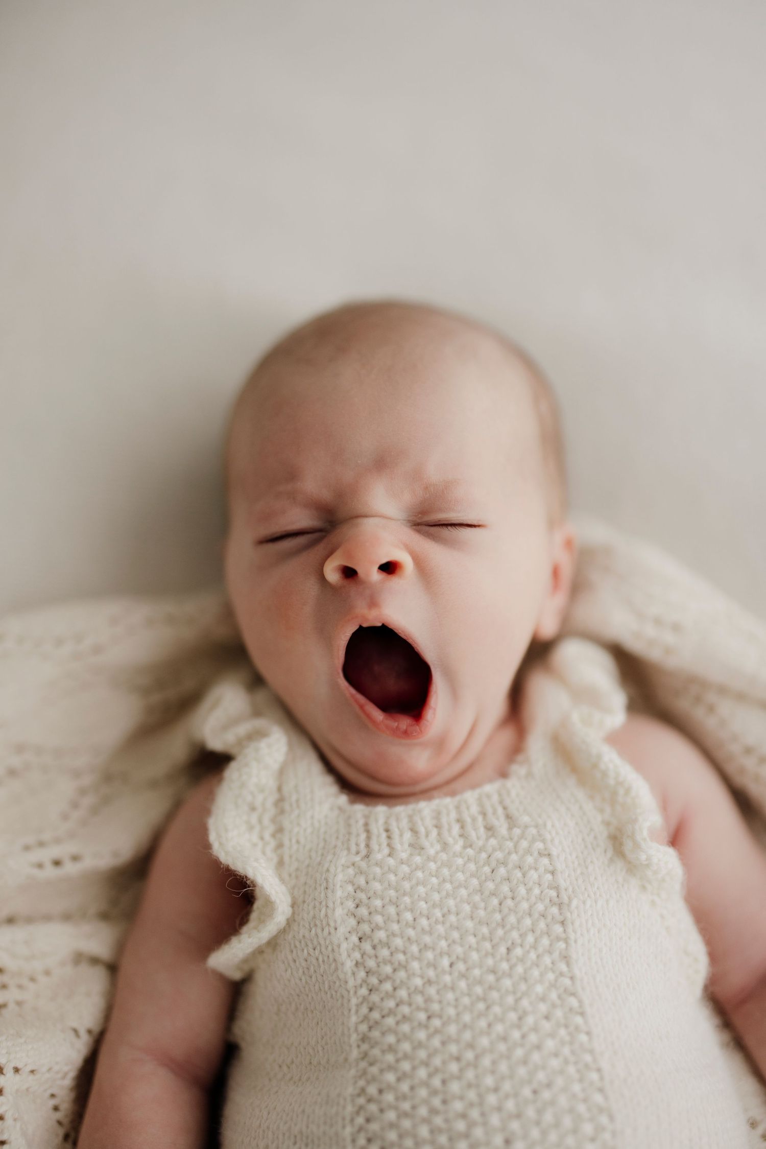 Adorable newborn baby in a cream knit outfit yawning widely while lying on a soft white blanket.