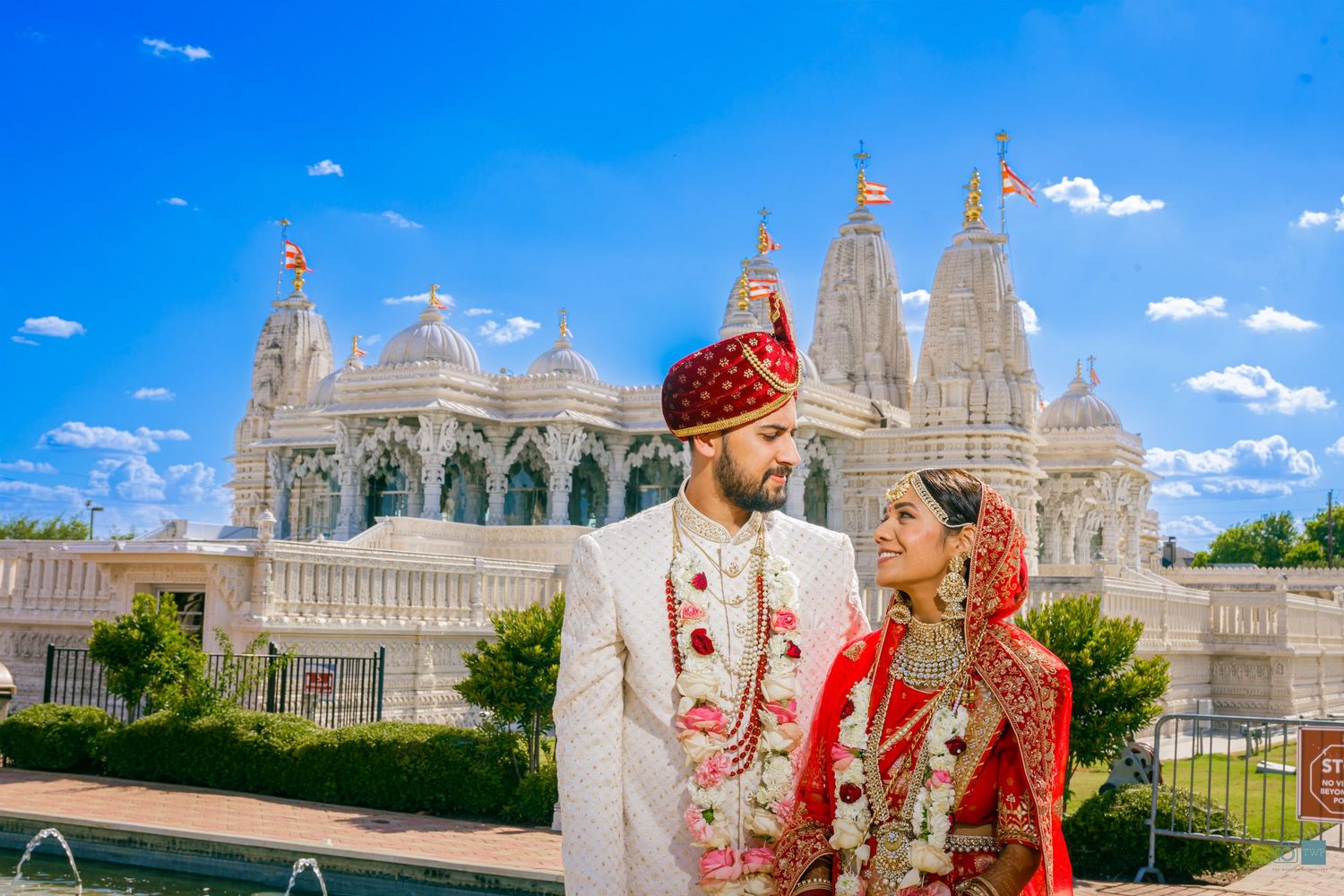 Bride and groom in traditional Indian wedding attire with floral garlands pose in front of an ornate white temple under a bright blue sky.
