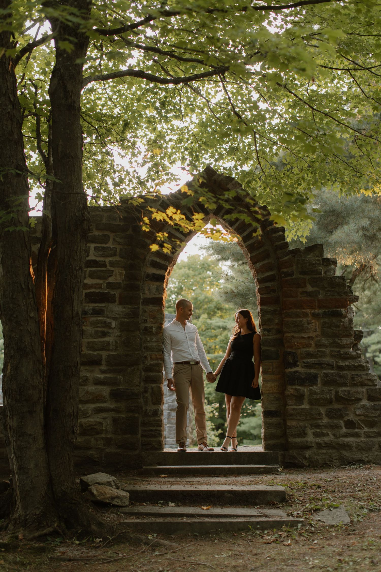 A couple stands on a bridge over a pond beneath dramatic weeping willow trees at sunset.