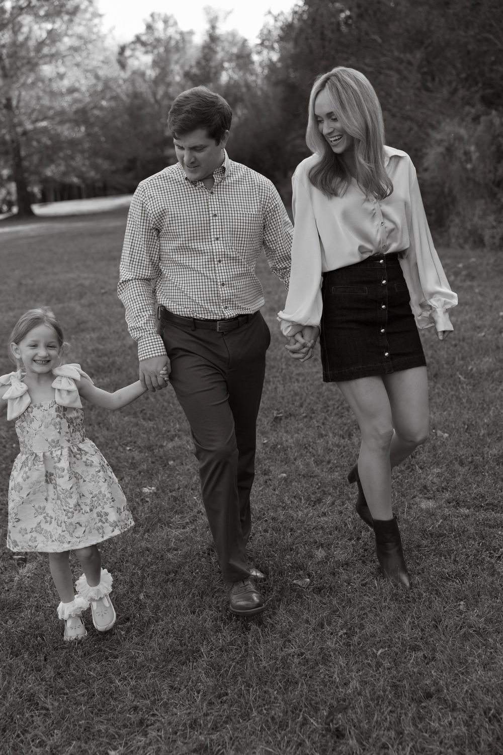 Black and white photo of a happy family of three walking outdoors, parents holding hands with young daughter in a floral dress.