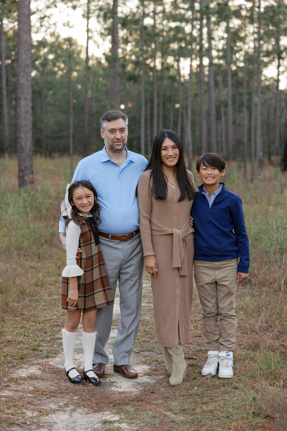 Family photo session outdoors with pine trees in the background and sandy ground in autumn.