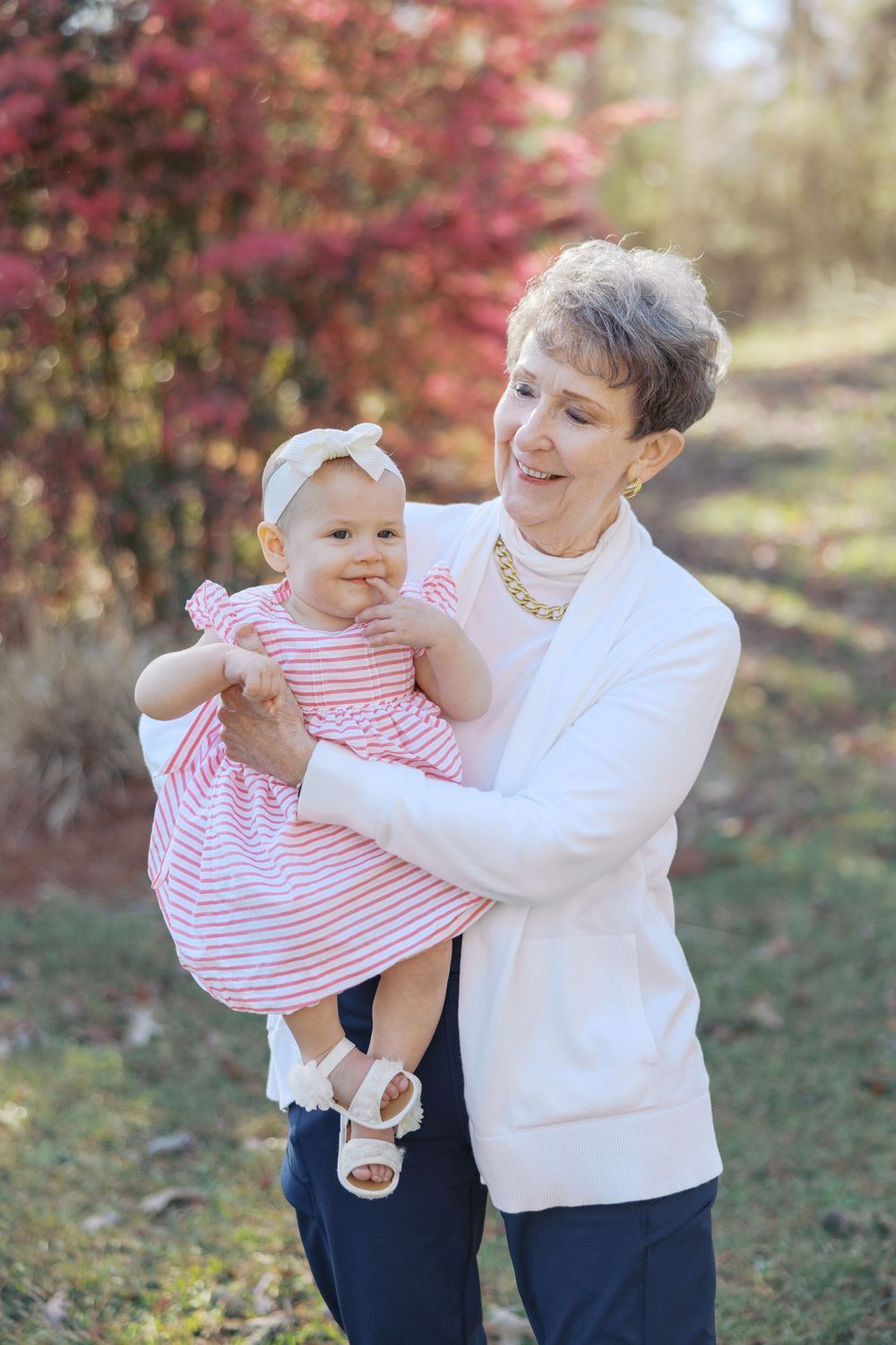 An adult in white sweater lifts and plays with a small child wearing red and white stripes in an autumn setting.