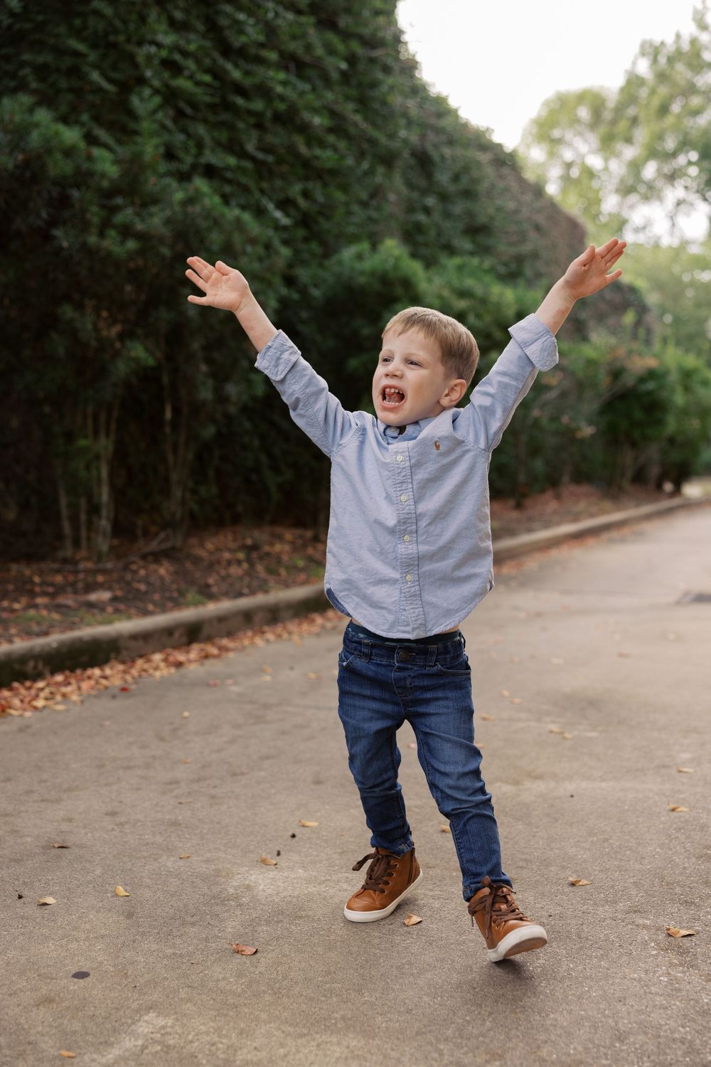 A young child in jeans and blue shirt joyfully jumps and plays on a tree-lined path covered with fallen leaves.