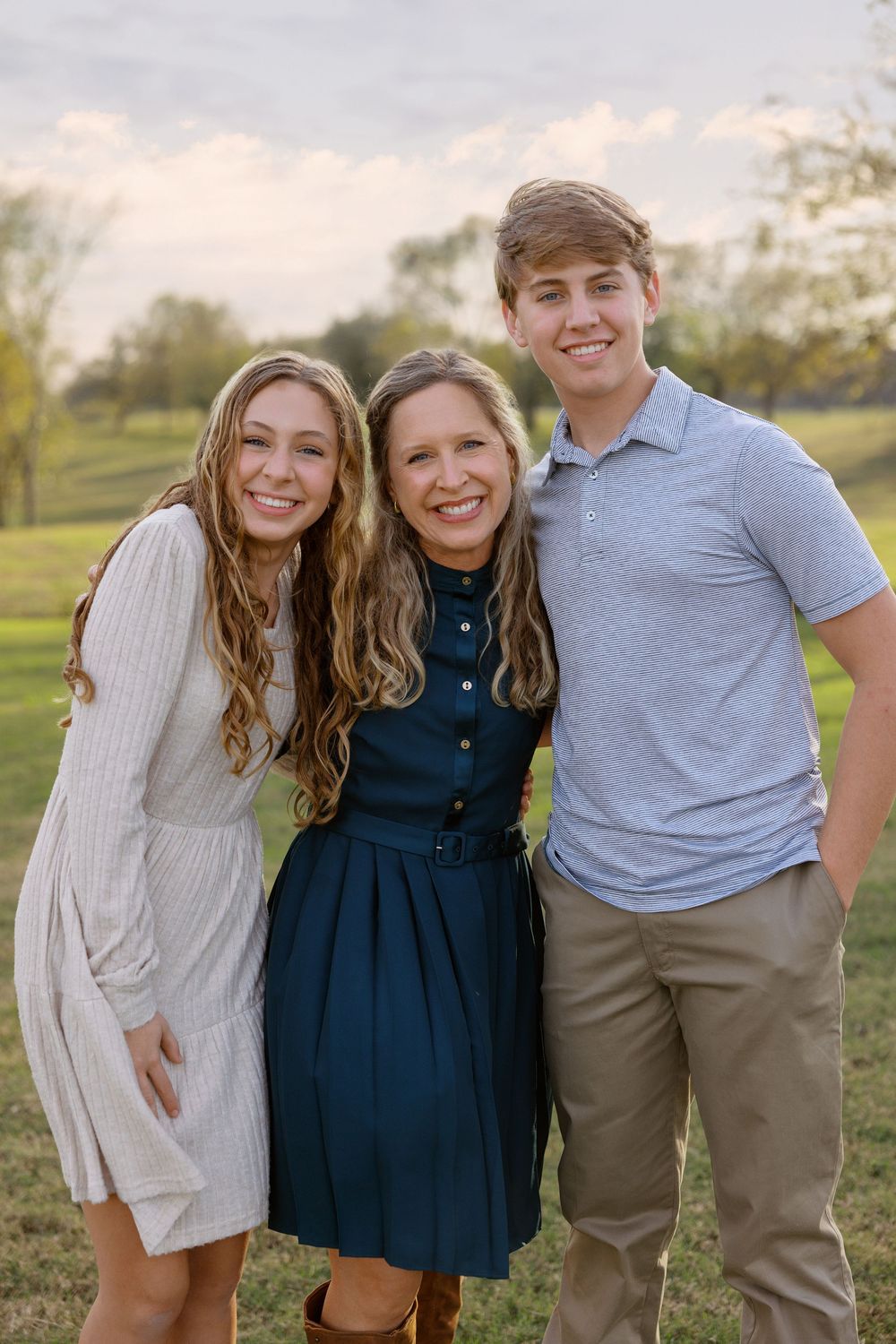 Three siblings posing together outdoors while wearing casual clothing on a sunny day in a park setting.