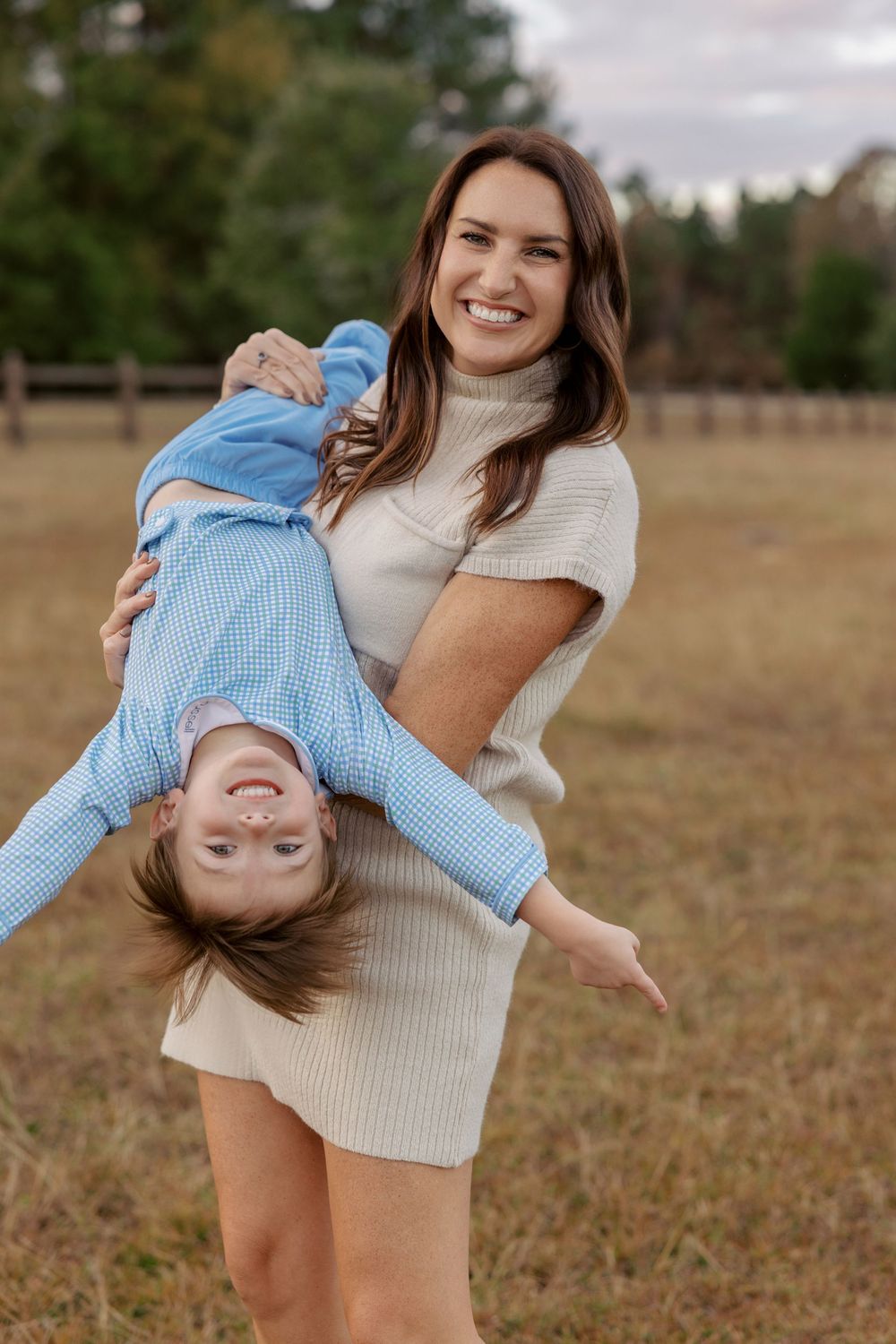 A smiling woman in a cream sweater dress playfully holds a laughing child upside down in an open field.