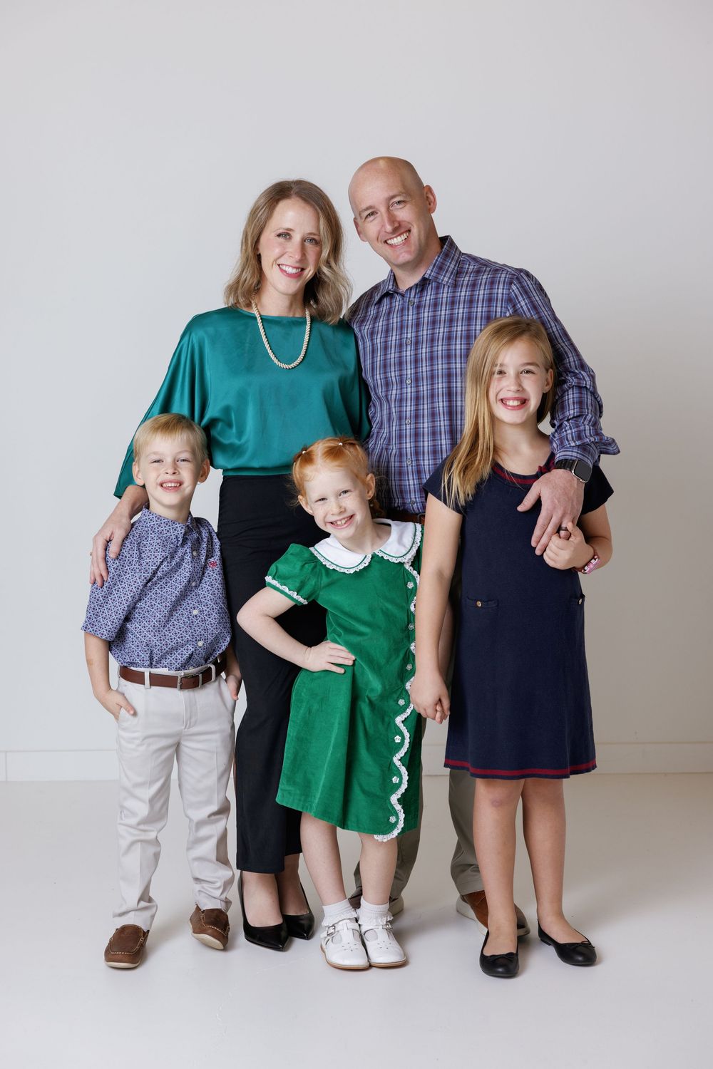 Family wearing coordinated green and navy blue outfits poses for professional portrait session against white backdrop.