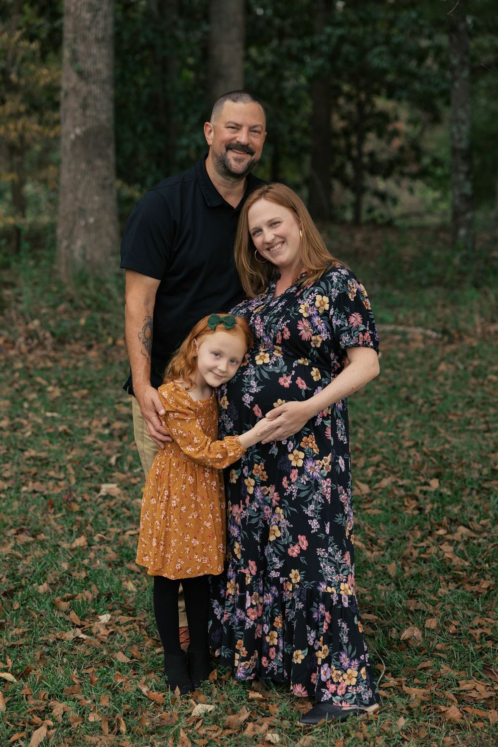 A family poses together in a wooded setting during autumn with fallen leaves on the ground.