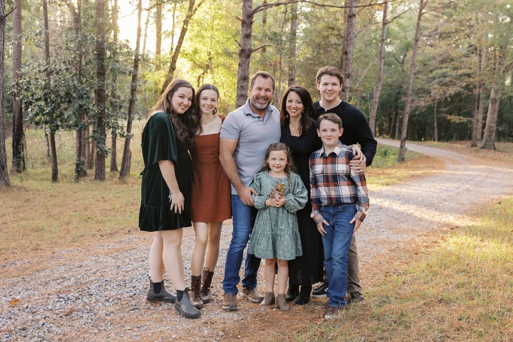 A group of people stand together outdoors on a wooded trail covered in autumn leaves.
