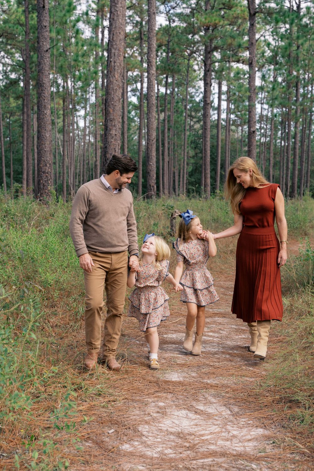 Family of four walking on a forest path among tall pine trees, wearing coordinated fall outfits in warm earth tones.
