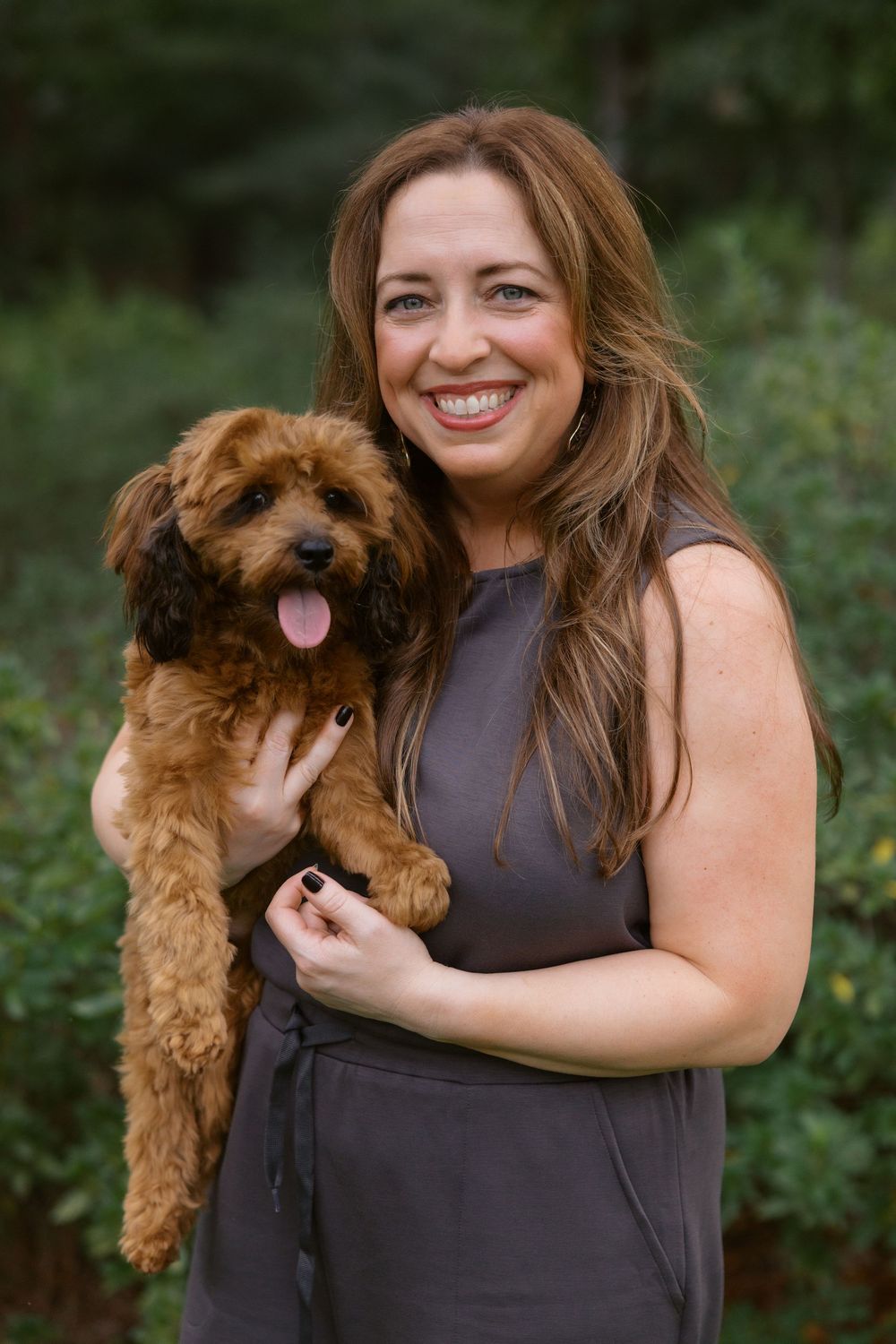 A person in a gray sleeveless dress holds a smiling brown Cavapoo dog outdoors against a natural green background.