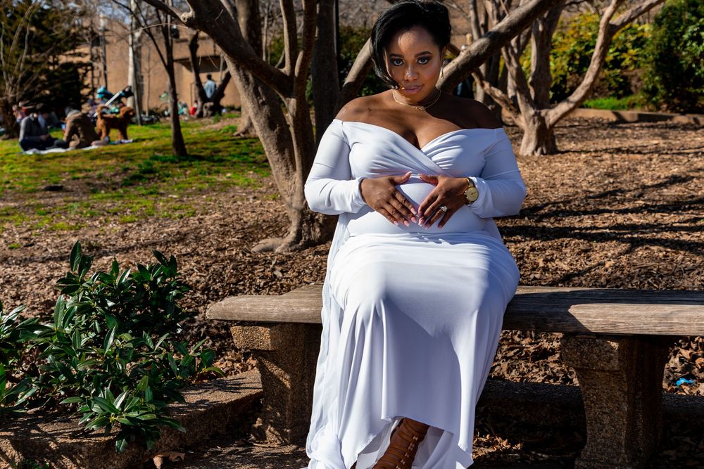 A pregnant woman in a flowing white dress sits on a stone bench in a tranquil garden setting during springtime.