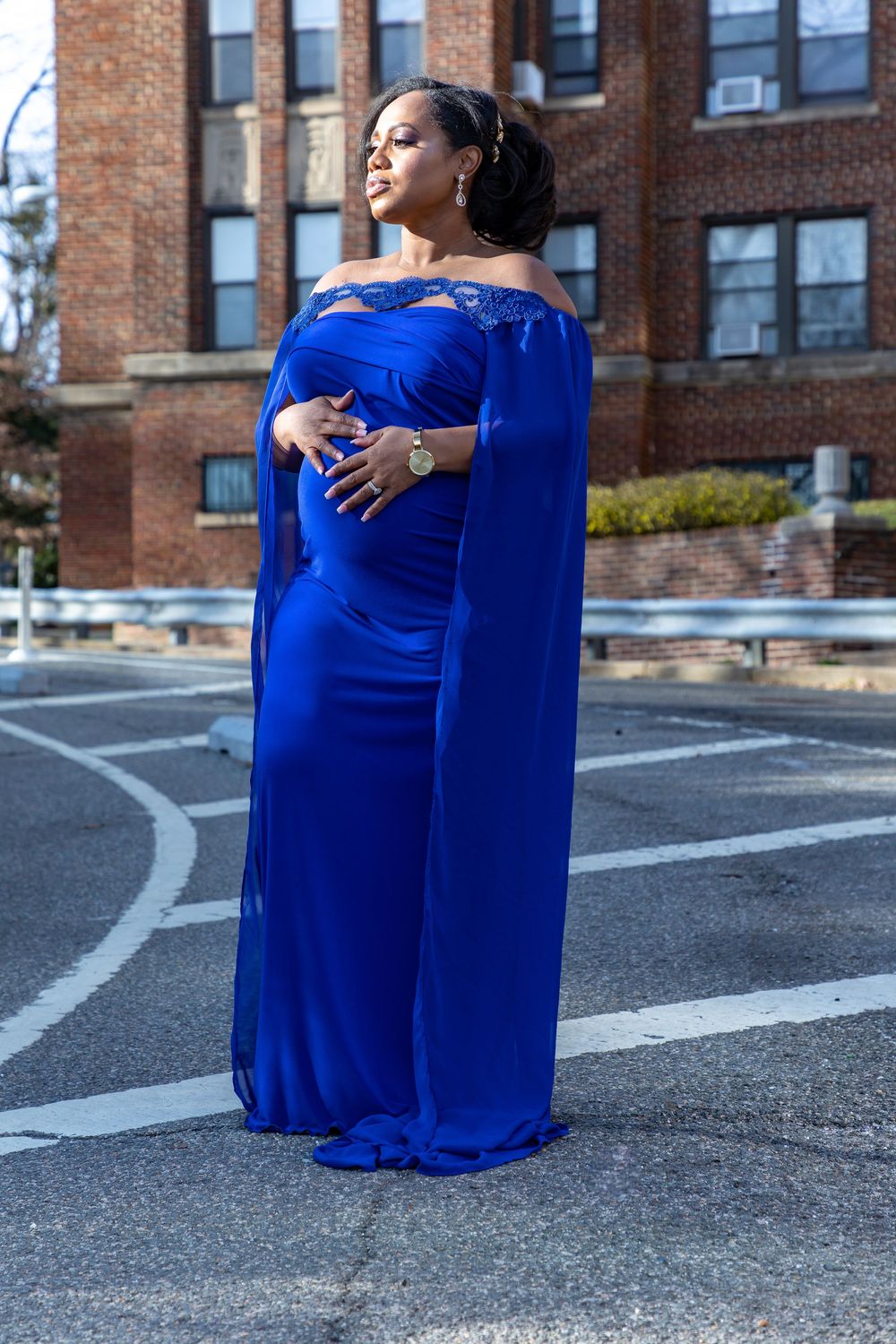 A figure in an elegant royal blue off-shoulder dress with a dramatic cape poses outdoors against a brick building.