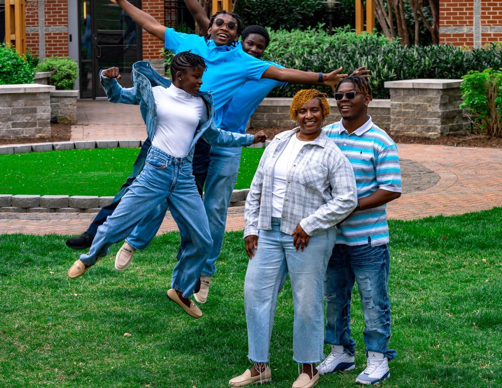 A group stands together on a grassy lawn in front of a brick building while two people jump playfully in the air.