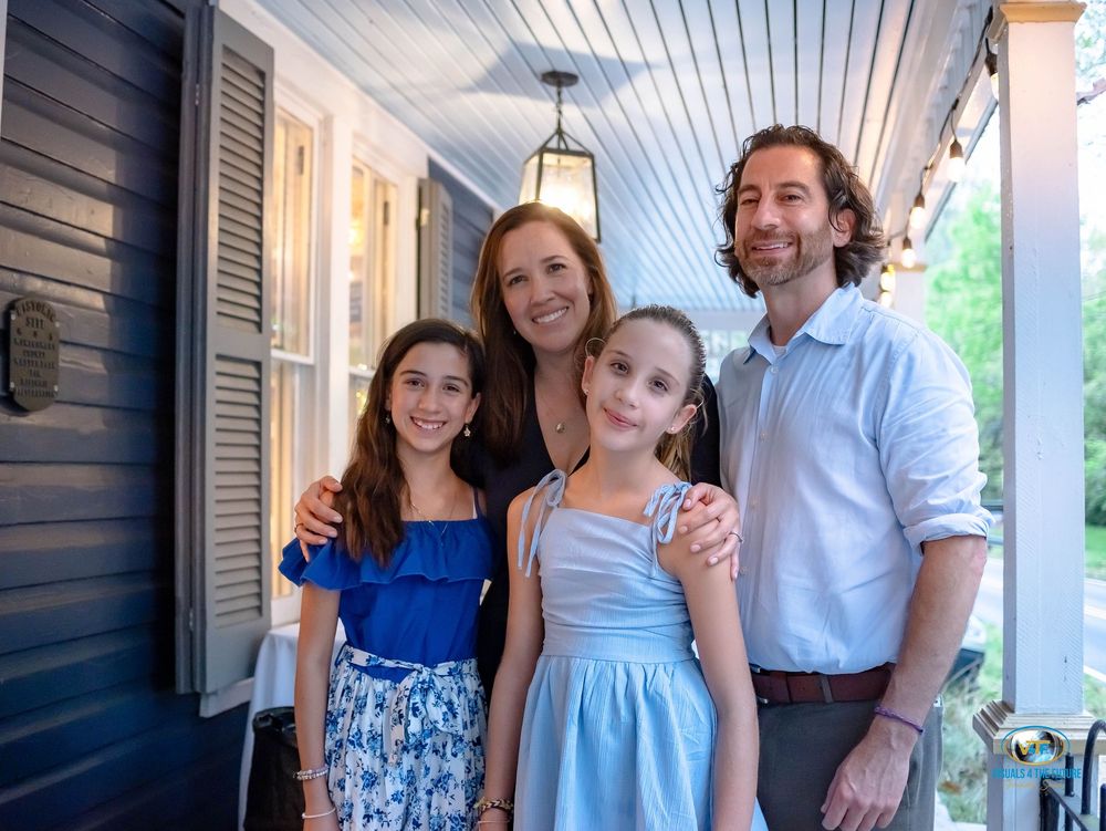 A family stands together on a porch with blue shutters and white columns on a sunny afternoon.