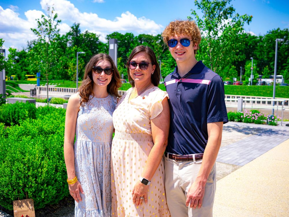 Three people wearing sunglasses pose together outdoors on a sunny day with lush greenery and a park in the background.