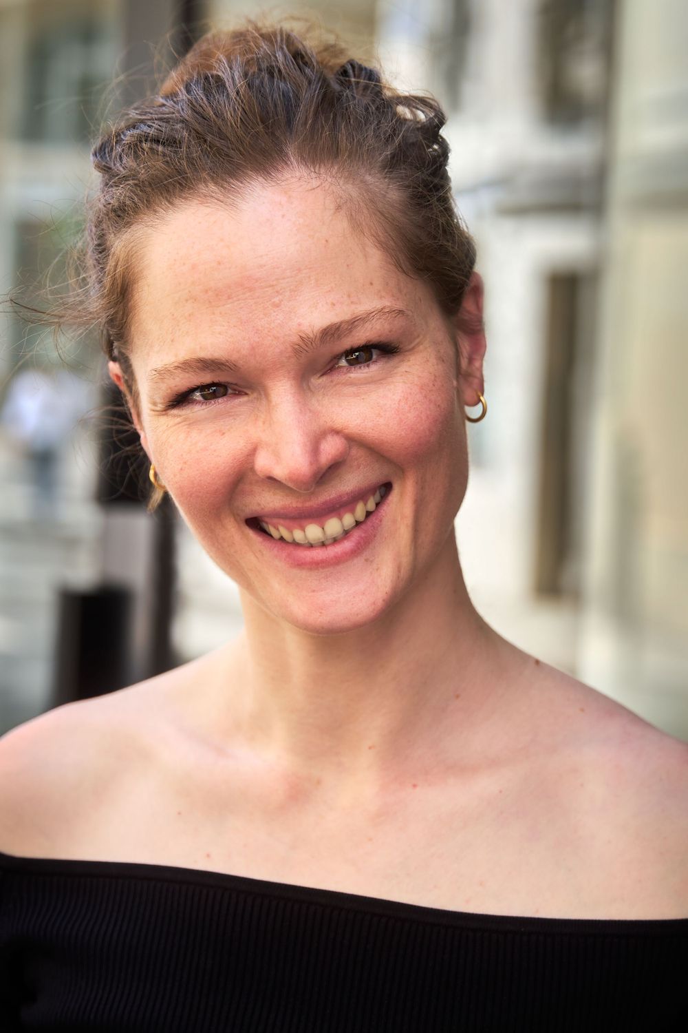 Portrait de femme en haut noir à épaules dénudées avec des cheveux relevés souriant chaleureusement.