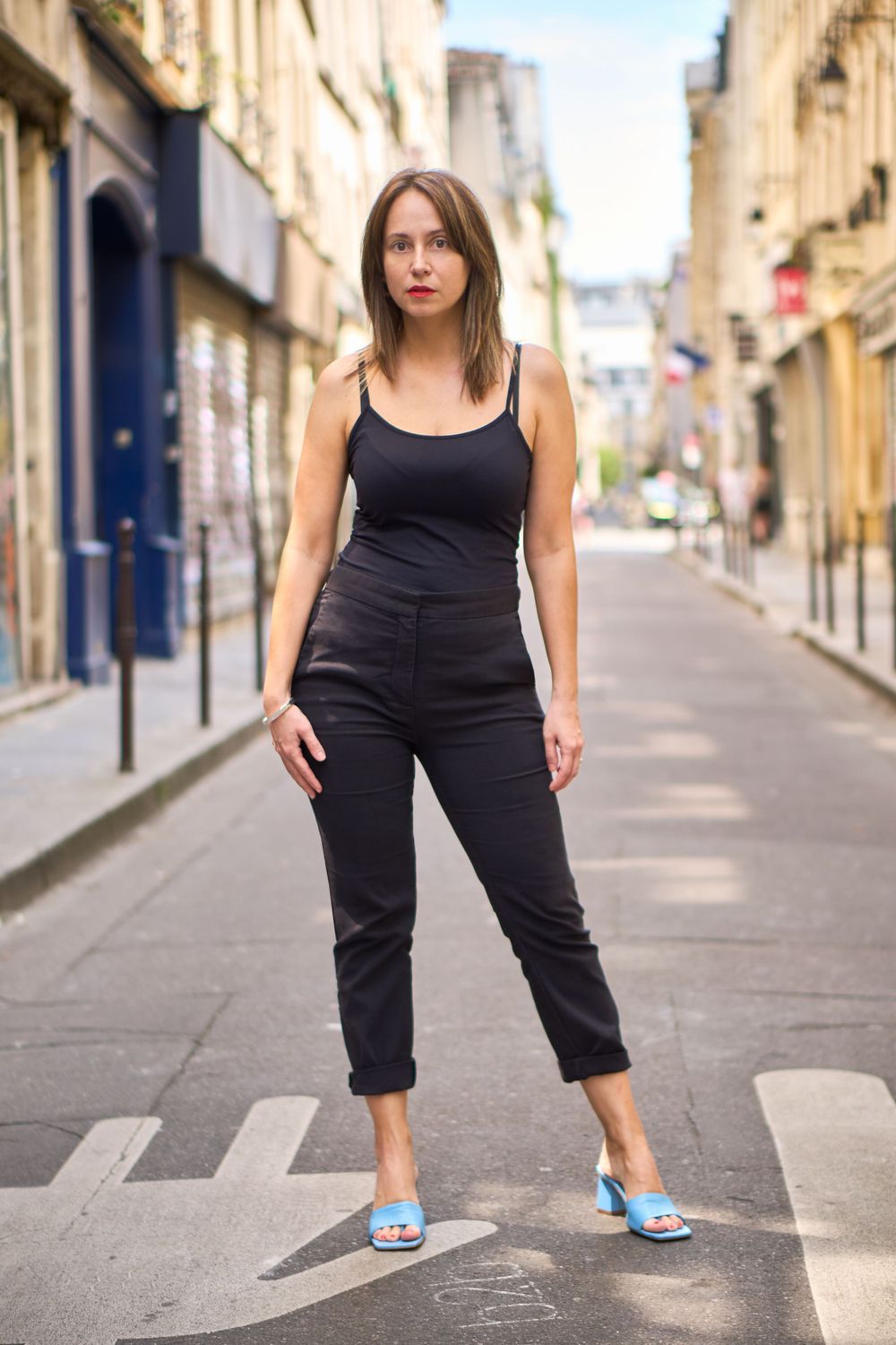 Photo de rue du corps entier en tenue noire, debout dans une rue pavée parisienne.