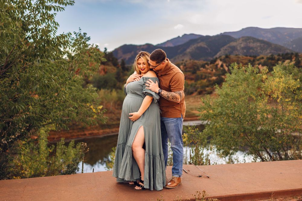 An expecting couple embraces while standing on a red rock formation with mountain views and autumn foliage in the background.