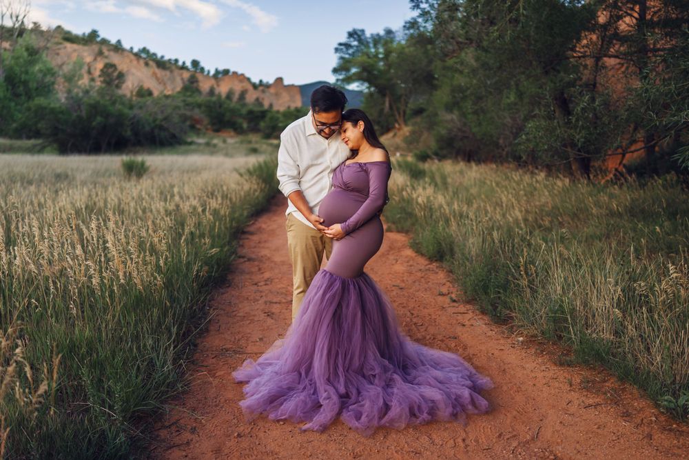 Couple poses on dirt path with flowing purple maternity gown during outdoor sunset shoot.