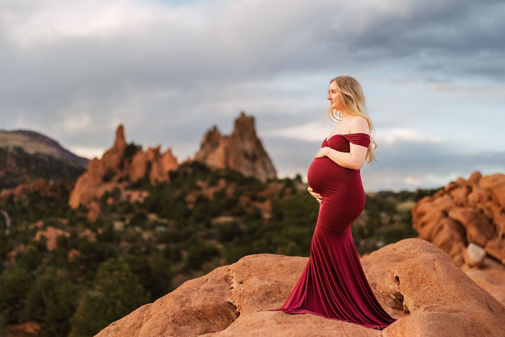 Pregnant woman in red off-shoulder gown poses on red rocks at Garden of the Gods, Colorado, with dramatic sky.