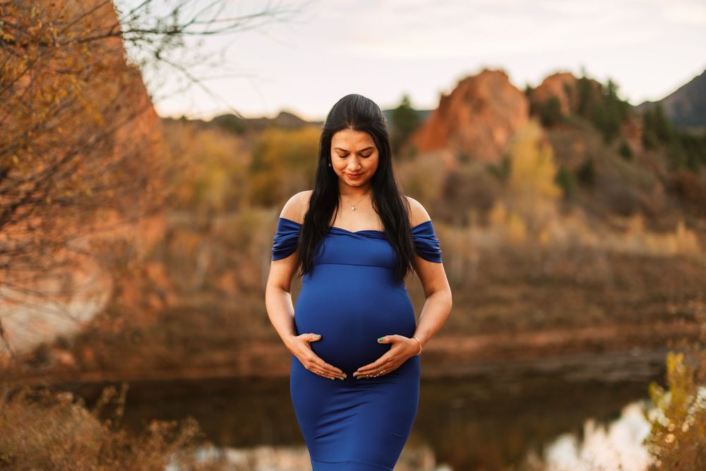 A pregnant woman in a fitted blue dress poses outdoors against a backdrop of autumn foliage and water in Colorado Springs, Colorado.