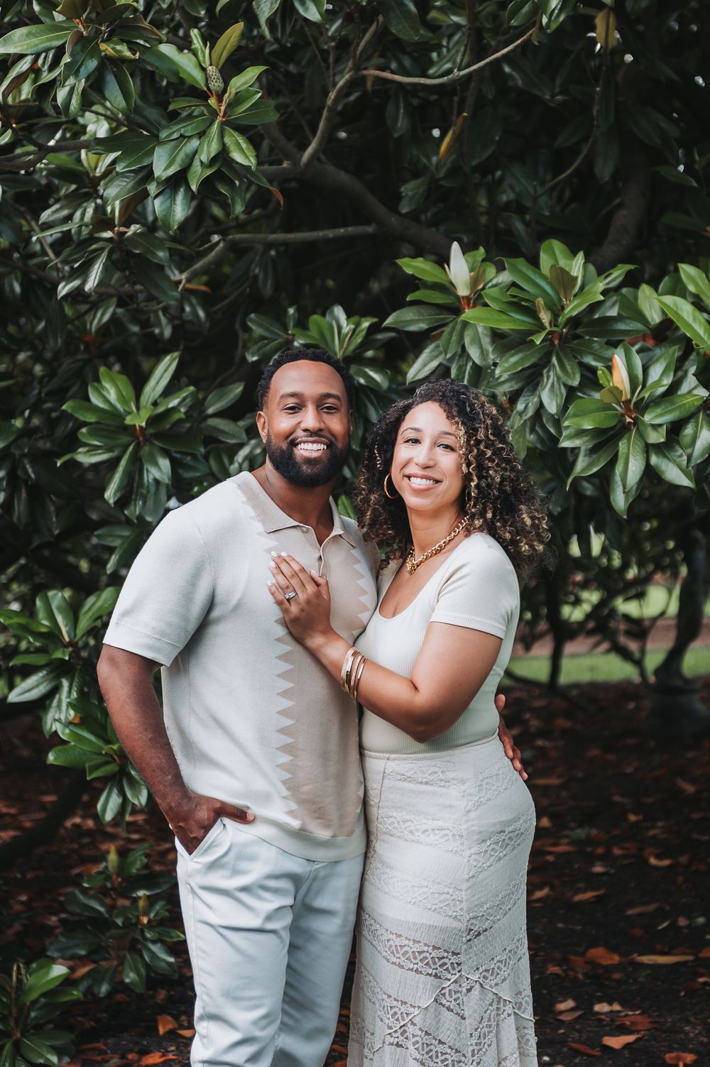 Joyful couple in white outfits laughing together in front of magnolia trees.