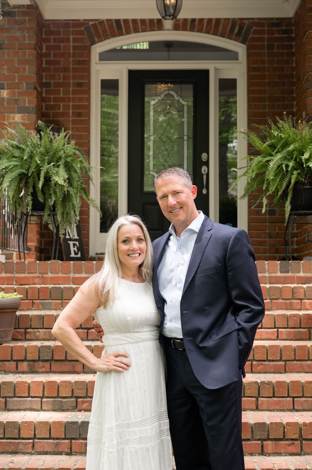 A couple poses together on a brick porch with decorative hanging plants framing the entrance.