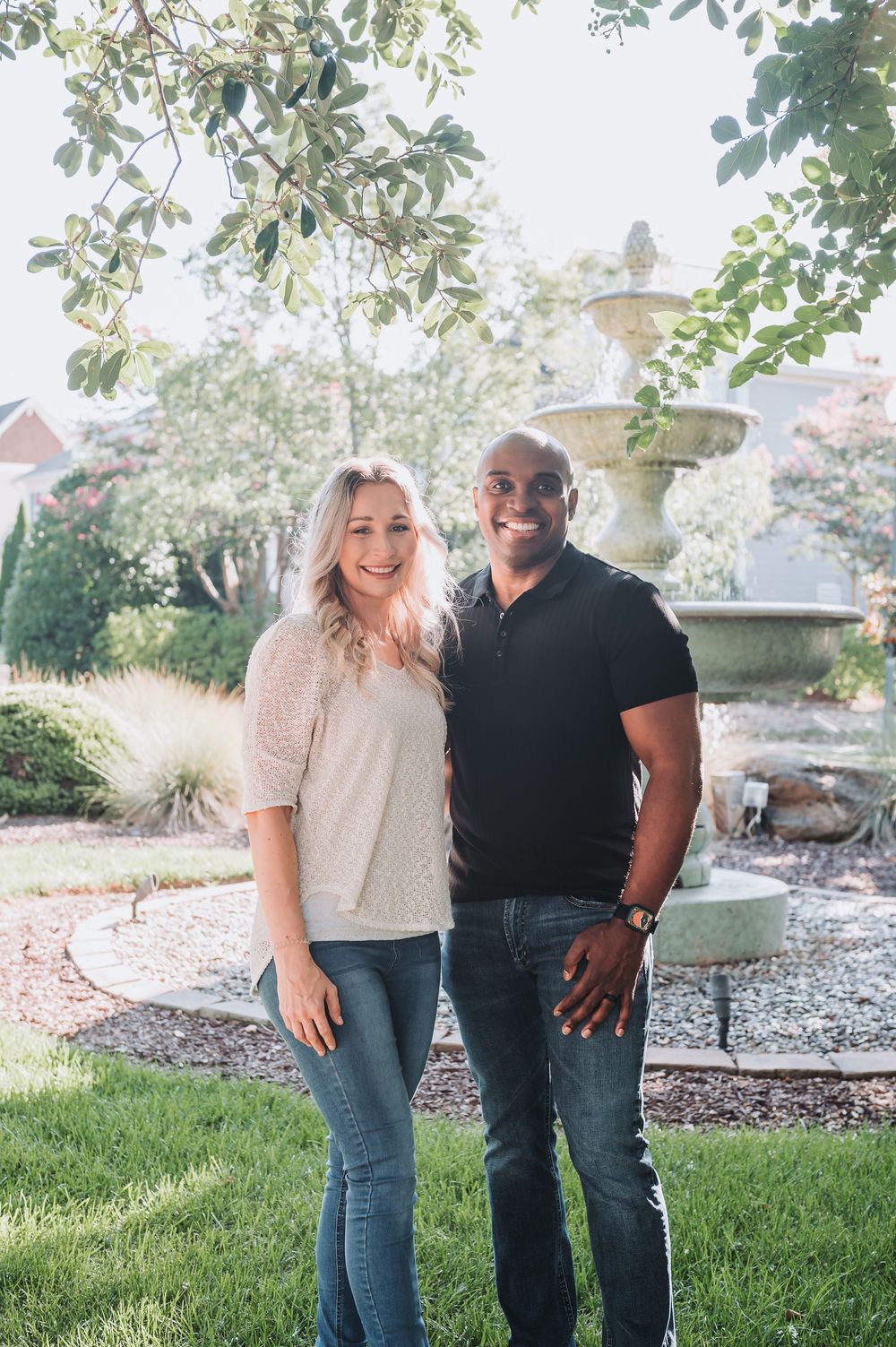 Casual couple poses together on grassy lawn with trees in background.