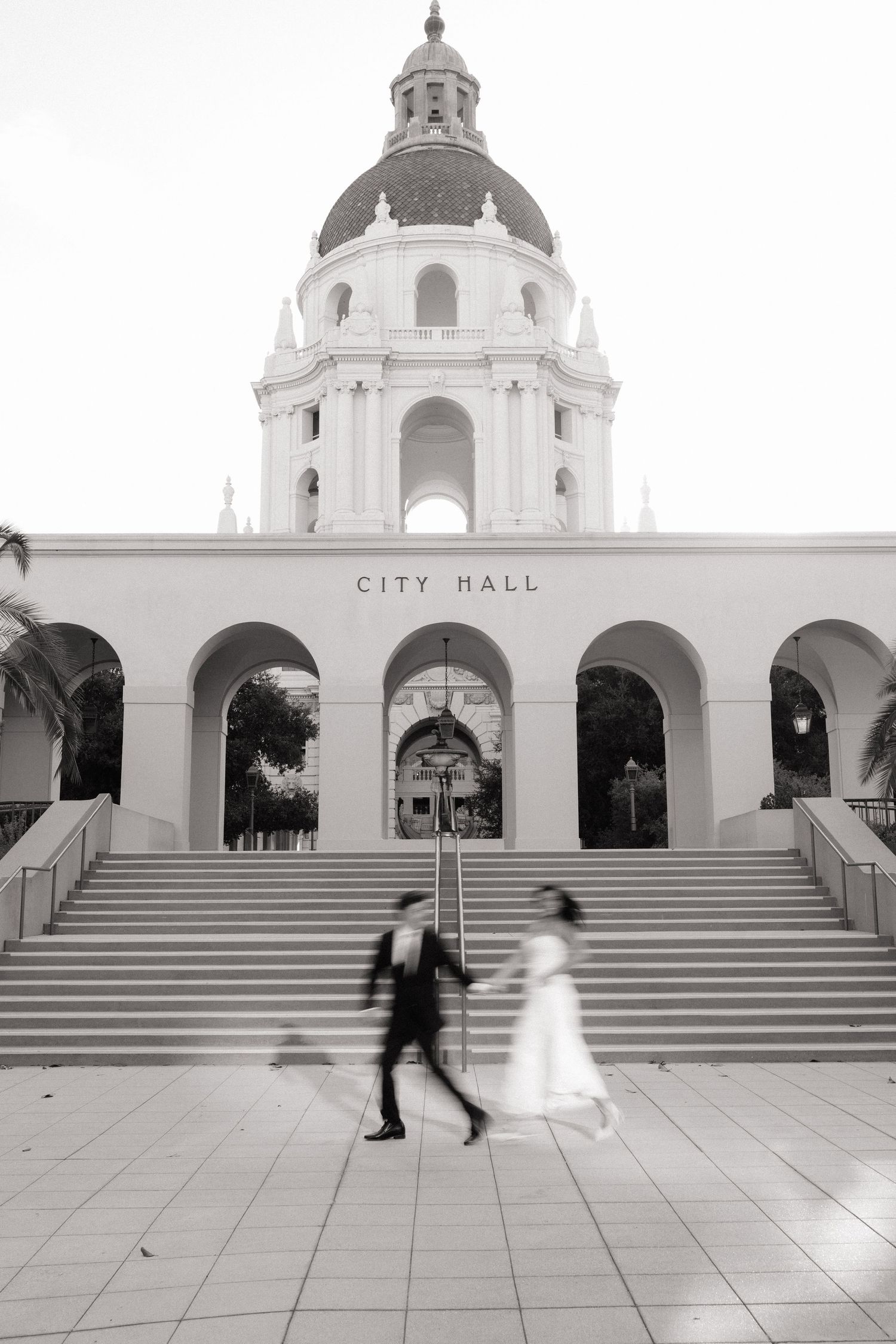 Black and white photo of a bride and groom walking past Pasadena City Hall steps. Fresno California Wedding Photographer and Videographer