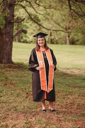 Graduate in black cap and gown with orange stole poses outdoors among trees.