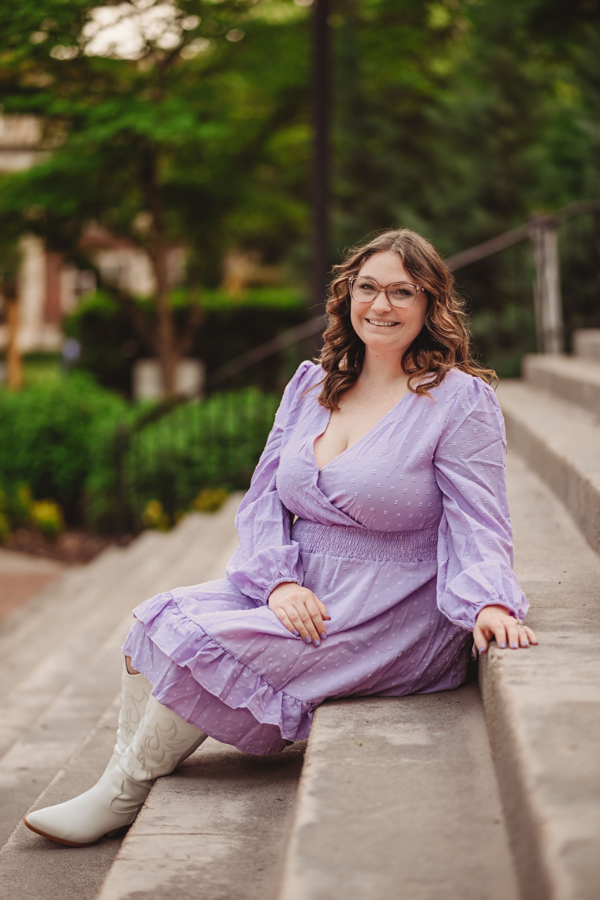 Graduate in a purple dress and boots poses on concrete steps during a photo session.