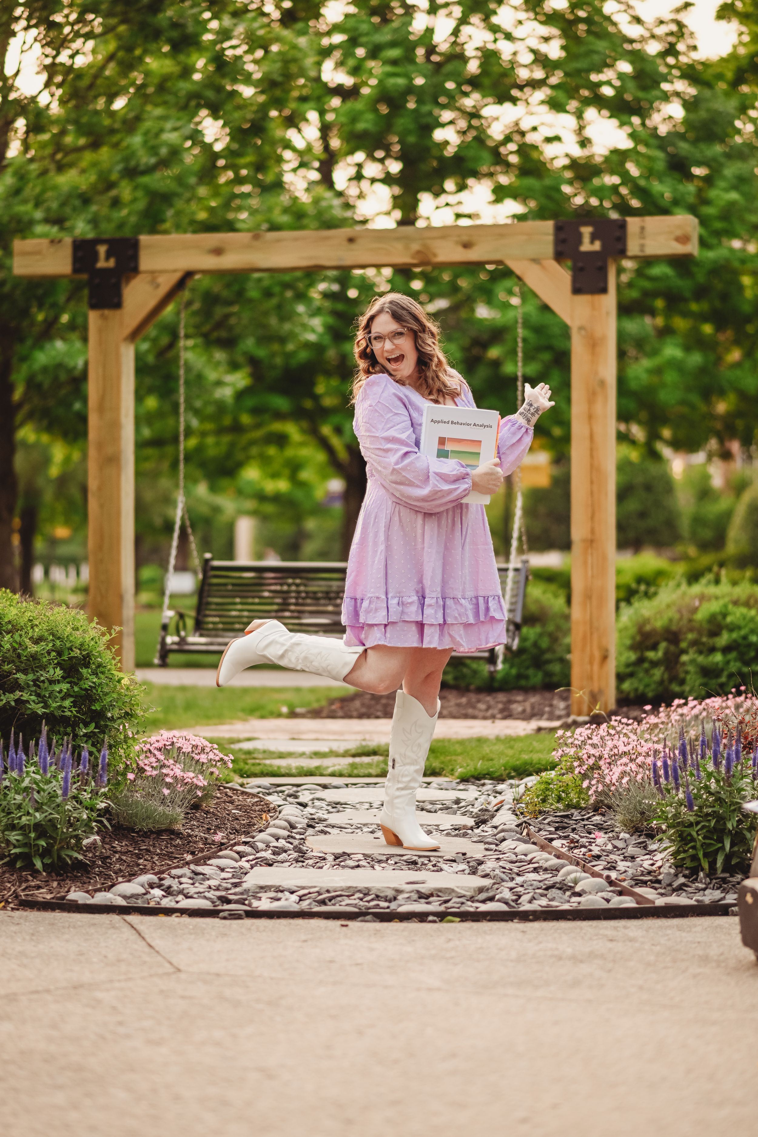 Graduate poses playfully on a garden path with wooden pergolas and purple flowers.