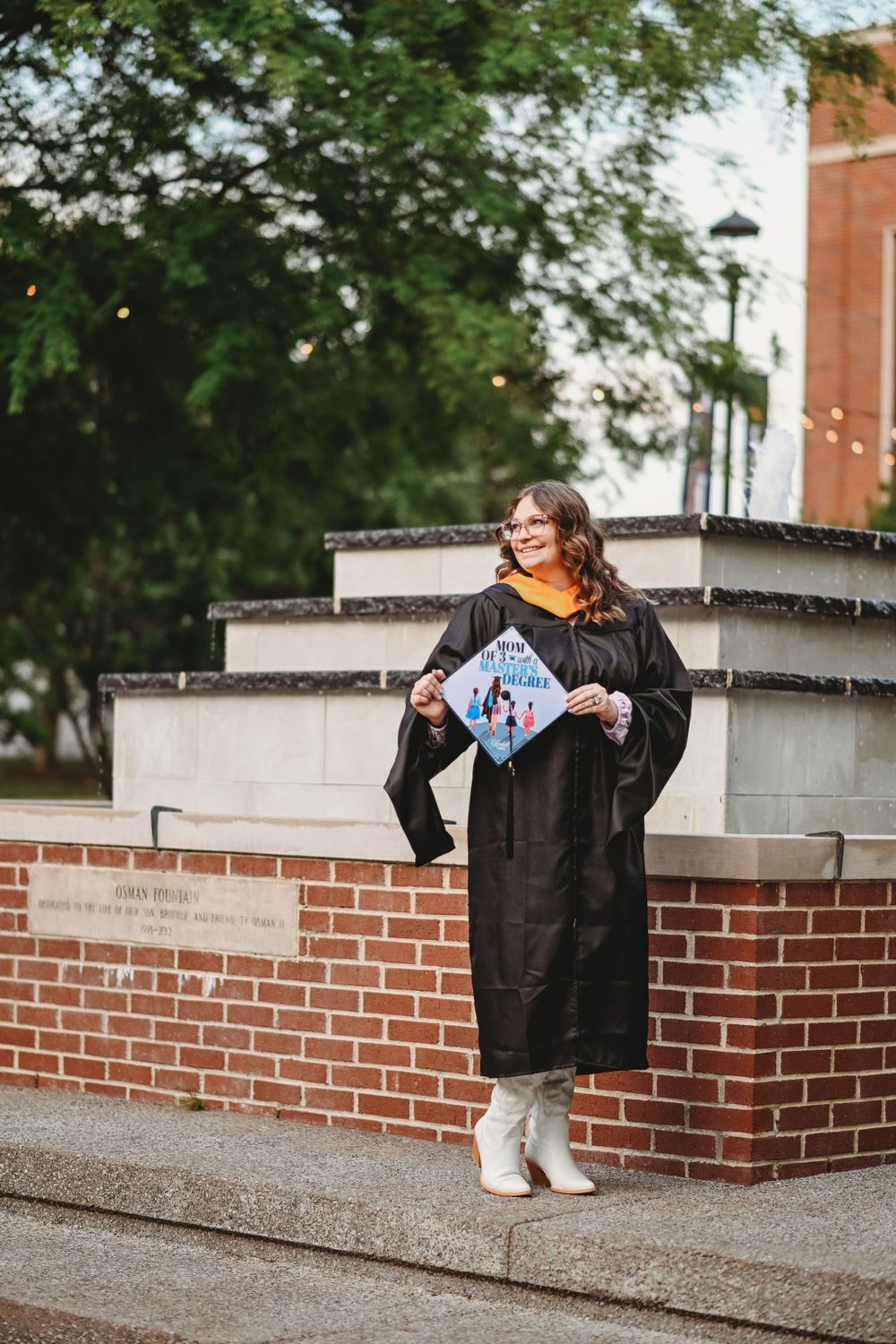 Graduate in black gown sits on brick wall during outdoor photo session.