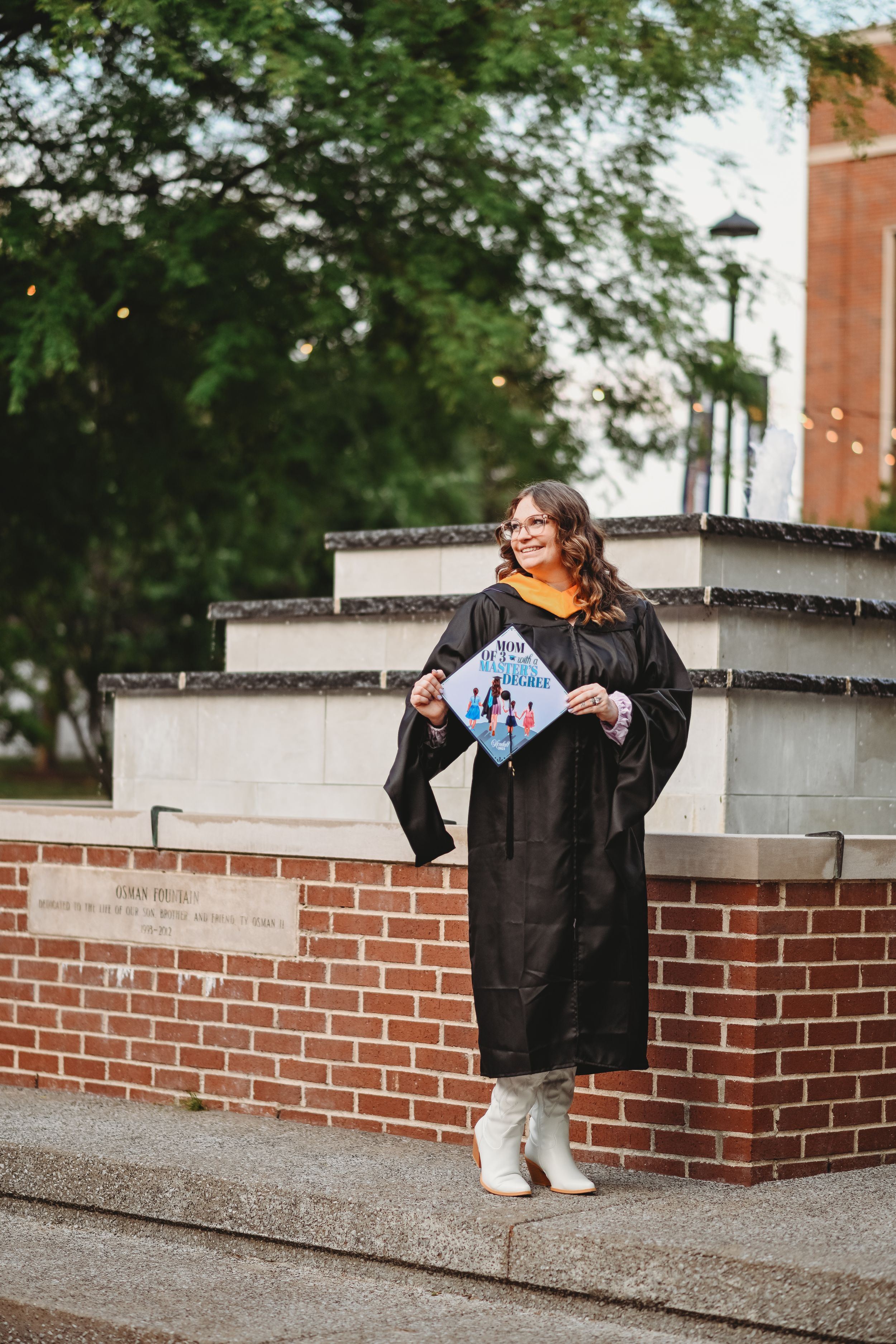 Graduate in black gown sits on brick wall during outdoor photo session.