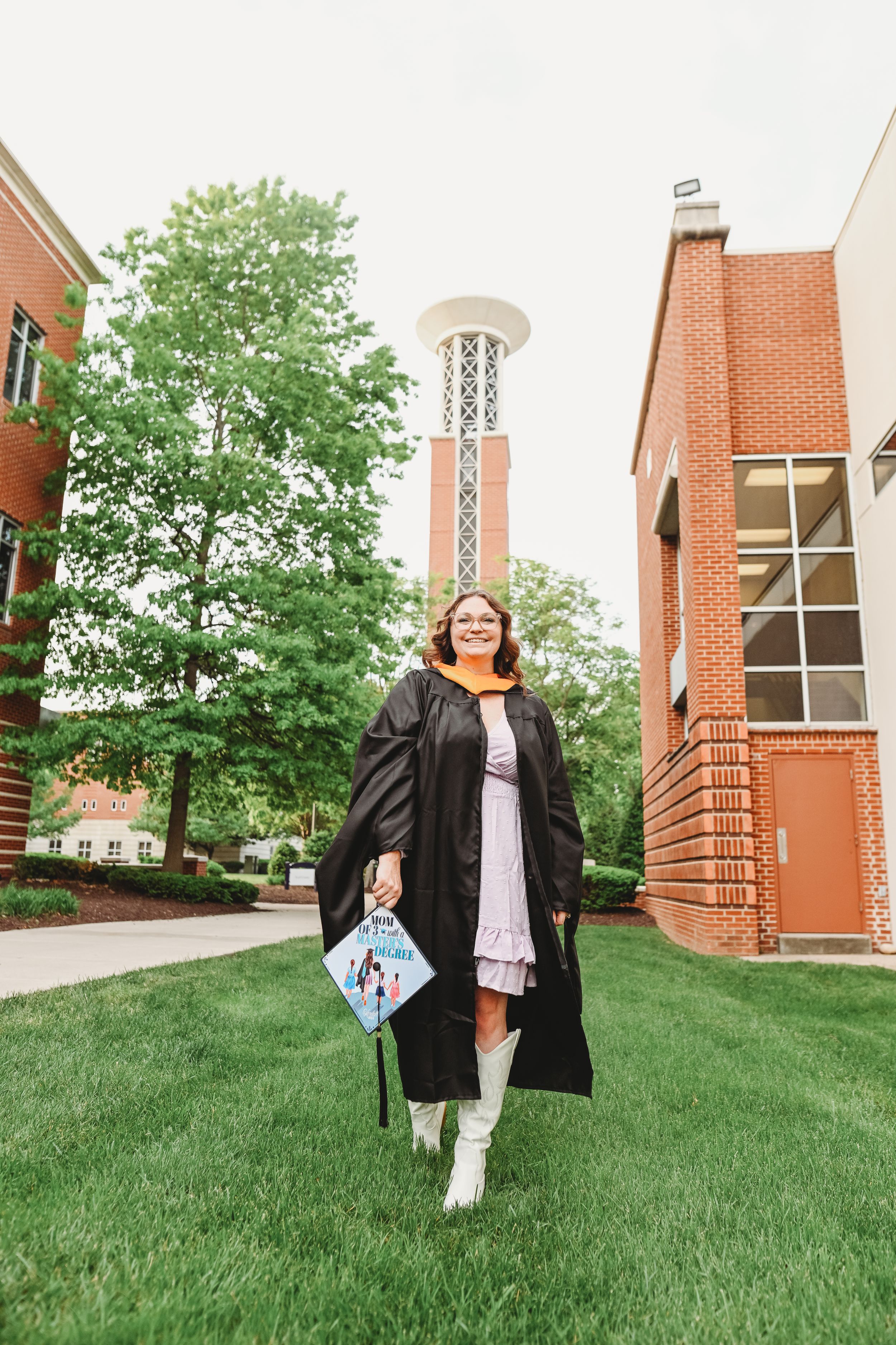 Graduate in black gown walks on grassy lawn near brick building.