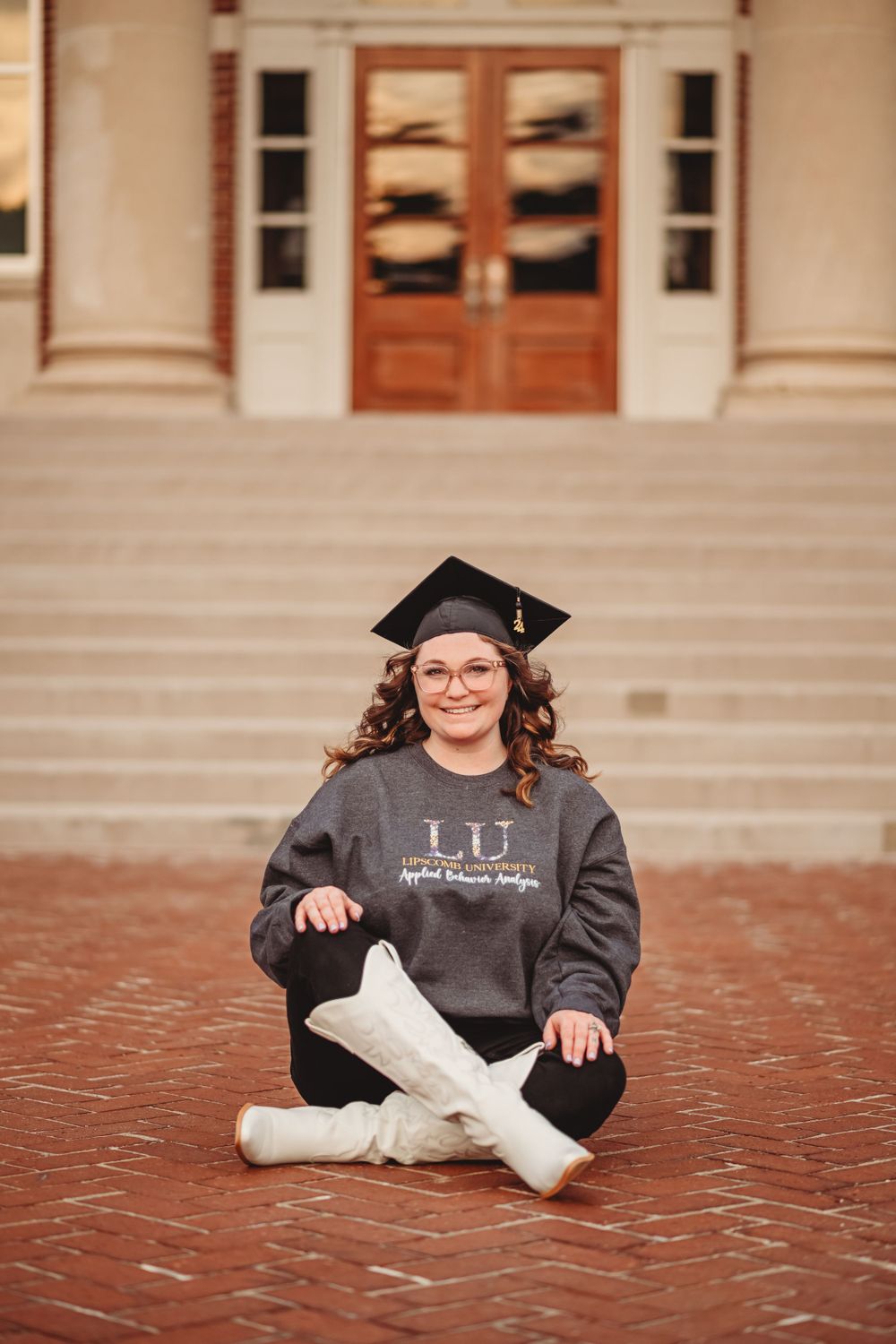 Graduate in black gown sits on red brick steps during graduation photoshoot.