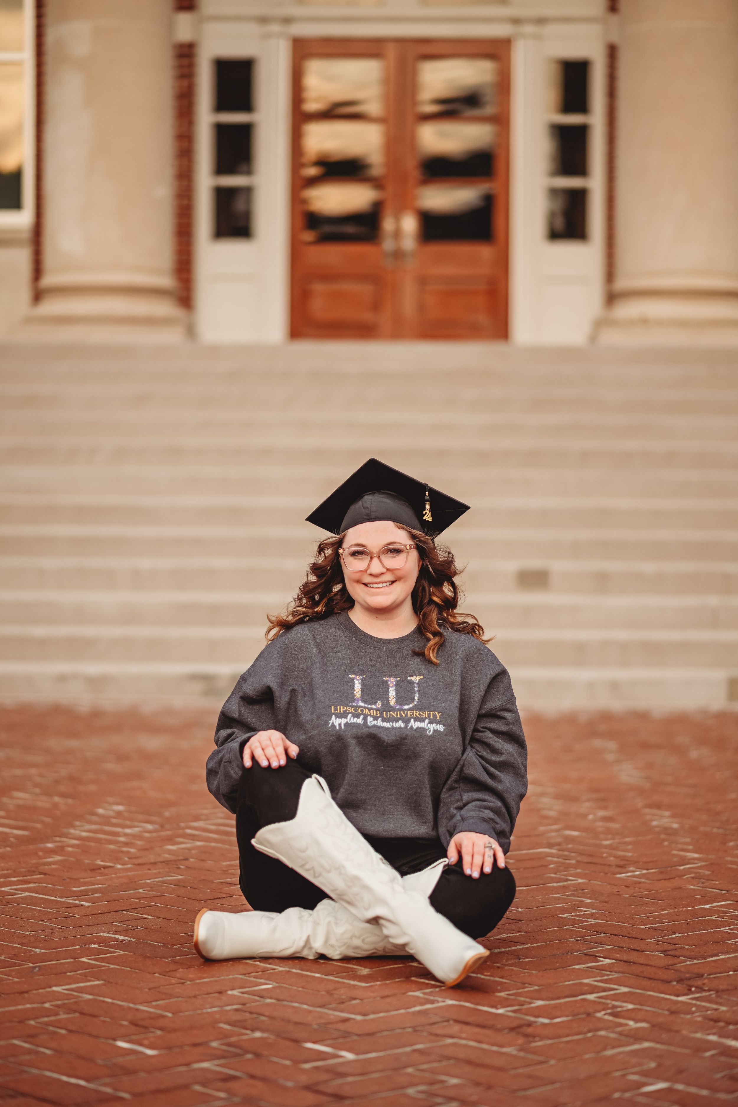 Graduate in black gown sits on red brick steps during graduation photoshoot.