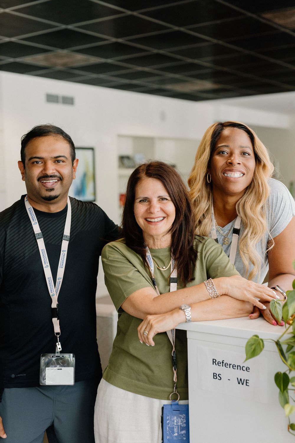 Three diverse colleagues smiling together near a reference shelf in a modern office workspace.