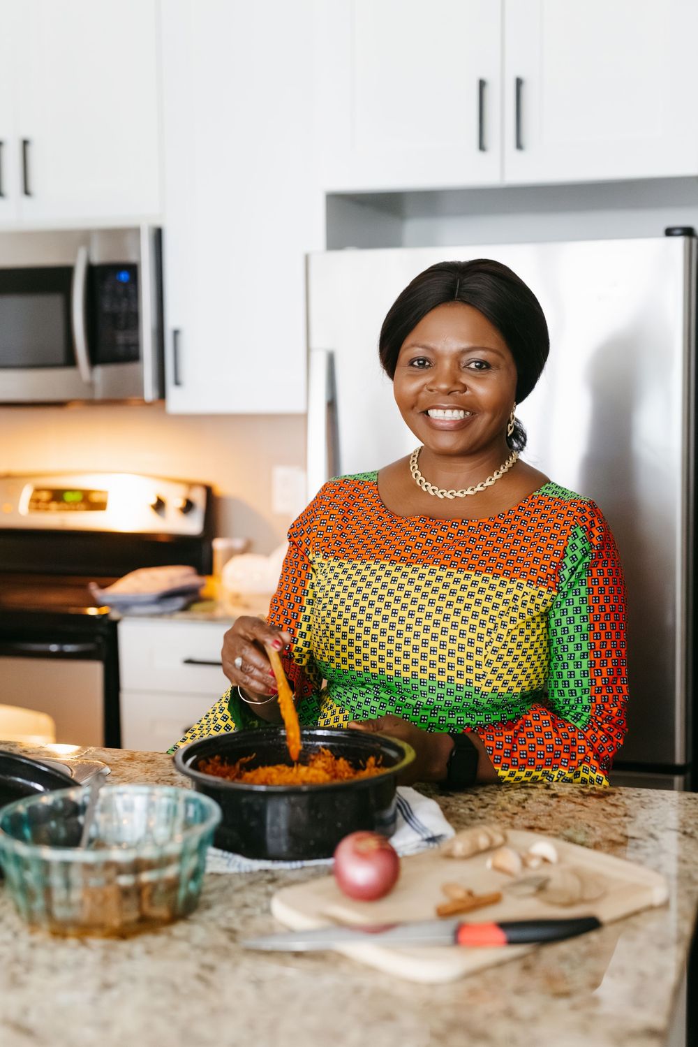 A smiling woman in colorful African print dress stirs food in a pot at a modern kitchen counter.
