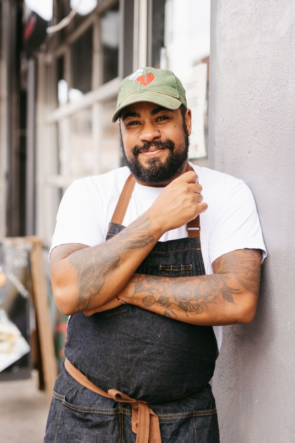 A tattooed male chef in a denim apron and green cap smiles while leaning against a wall outside.