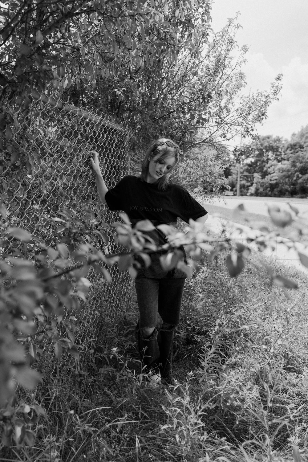 A young woman in black clothing leans against a chain-link fence surrounded by overgrown plants in black and white.