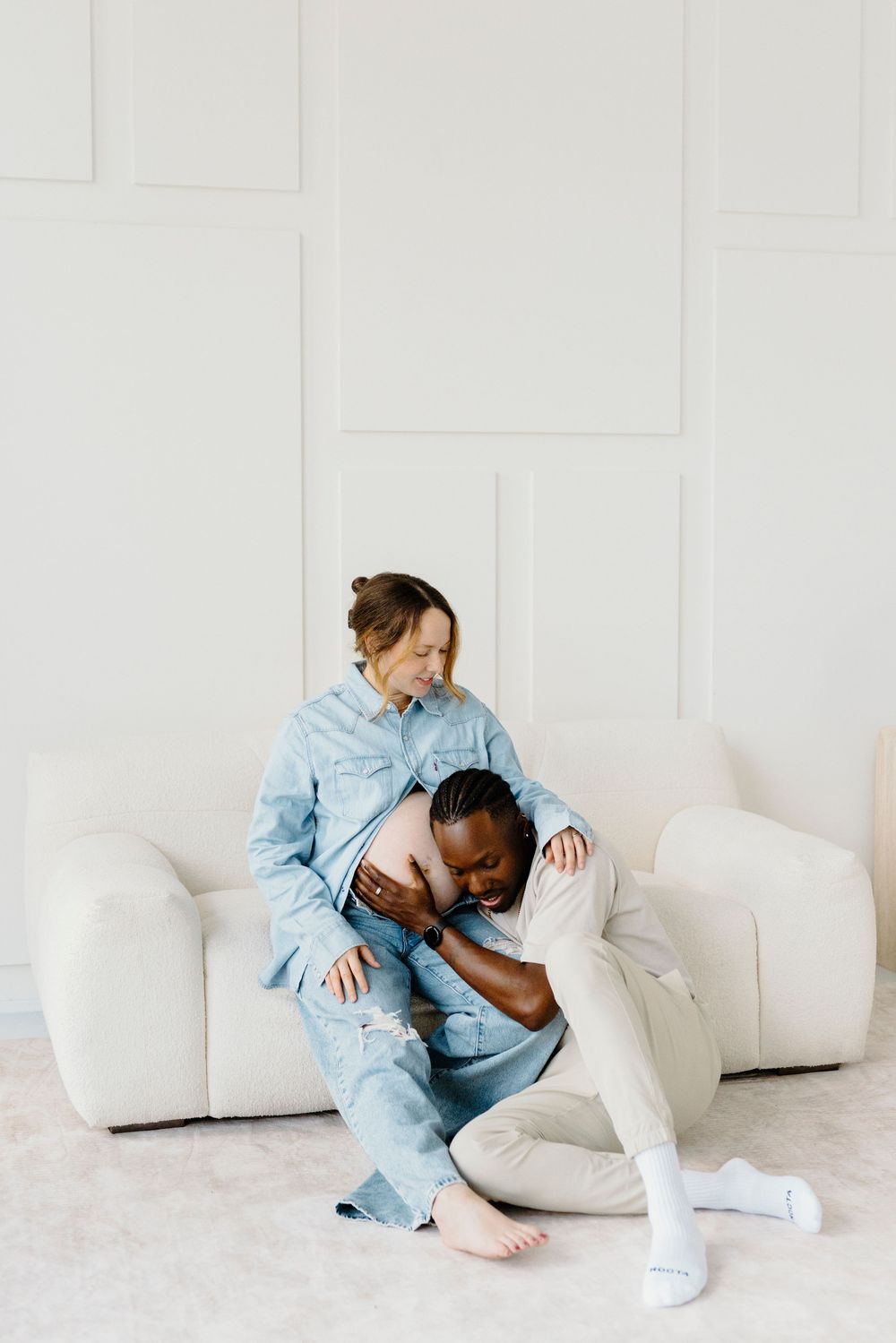 Interracial couple sharing a tender maternity moment on a white sofa, man kissing pregnant woman's belly.